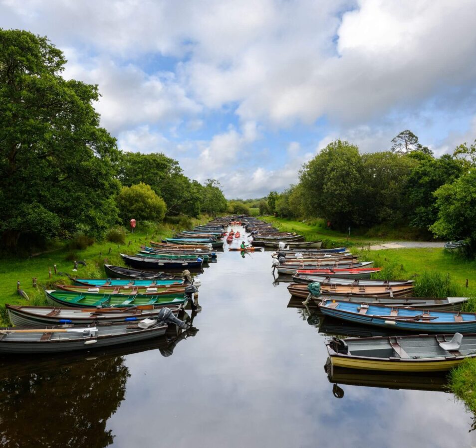 Families enjoying a serene day by boats on a lush river in Killarney, under a cloudy sky.