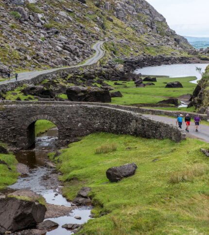 Family strolling on a scenic path near ancient stone bridge and lake, surrounded by lush greenery.