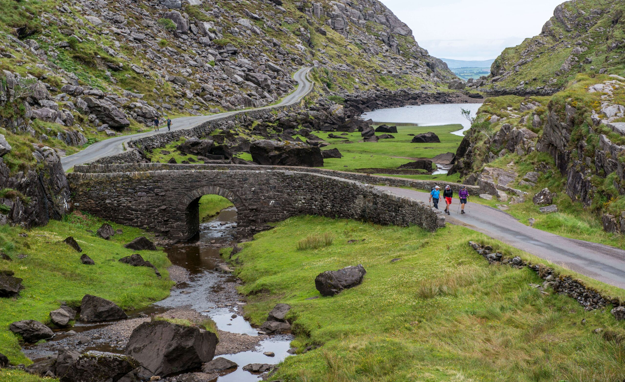 Family strolling on a scenic path near ancient stone bridge and lake, surrounded by lush greenery.