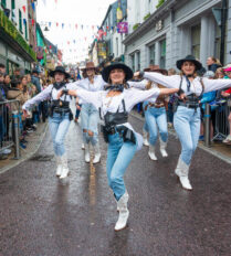 A lively parade with dancers in western attire performing in a vibrant Killarney street.