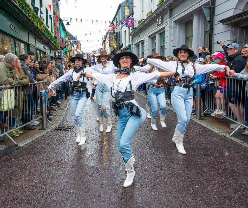 A lively parade with dancers in western attire performing in a vibrant Killarney street.