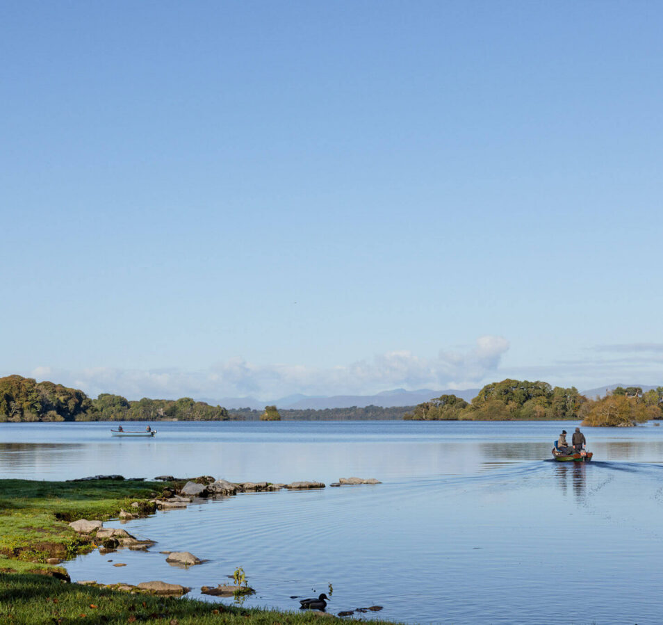 A family enjoys a serene boat ride on a calm lake, surrounded by lush greenery and distant hills.