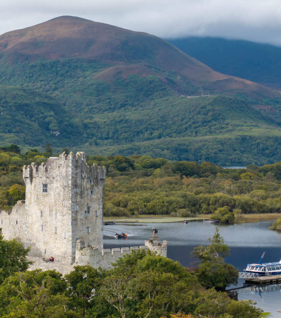 Scenic view of a castle by a lake with a boat, surrounded by lush greenery and mountains in Killarney.