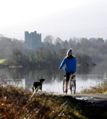 A person cycling with a dog beside a tranquil lake, with a castle in the background at Killarney.
