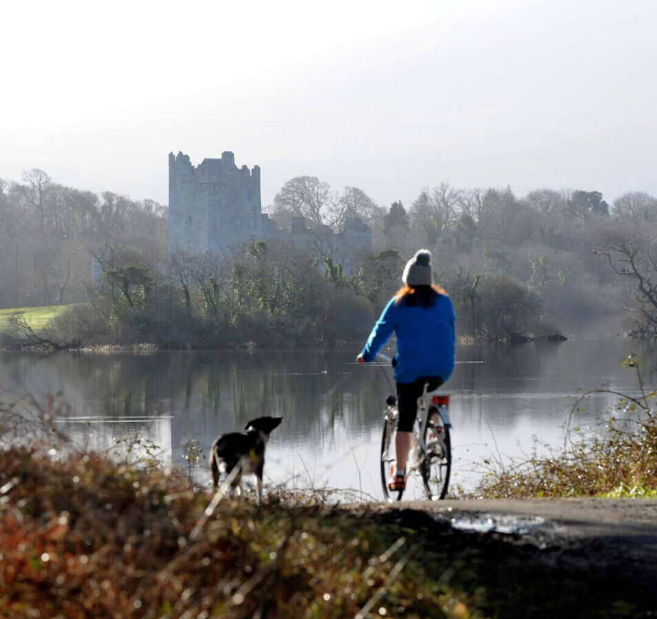 A person cycling with a dog beside a tranquil lake, with a castle in the background at Killarney.