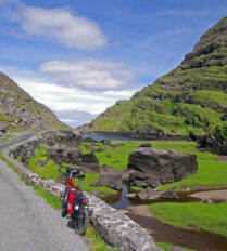A bicycle leans against a stone wall on a scenic road with lush green hills and a clear sky.