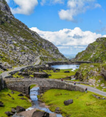 A family strolls on a stone bridge over a stream, surrounded by lush hills in Killarney, Ireland.