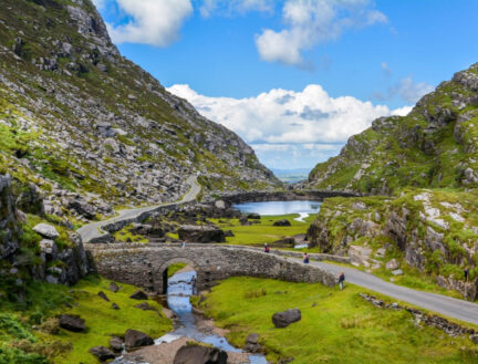 A family strolls on a stone bridge over a stream, surrounded by lush hills in Killarney, Ireland.