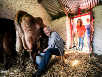 A man milking a cow in a rustic barn as a family watches, enjoying a farm tour.