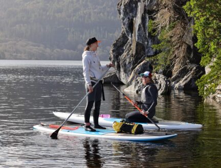 A couple paddleboards on a serene lake in Killarney, surrounded by lush greenery and rocky cliffs.