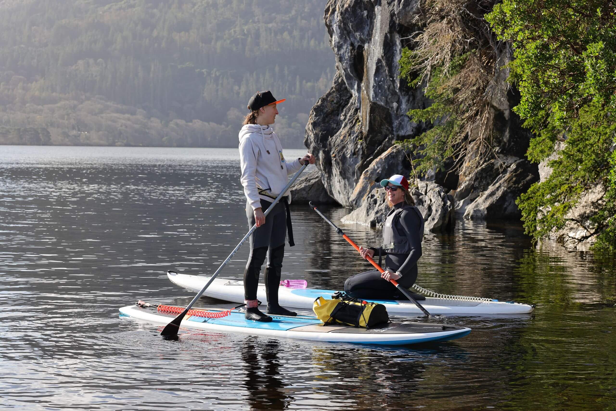 A couple paddleboards on a serene lake in Killarney, surrounded by lush greenery and rocky cliffs.