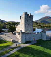 A couple strolls by a historic castle near a serene lake and mountains in Killarney.