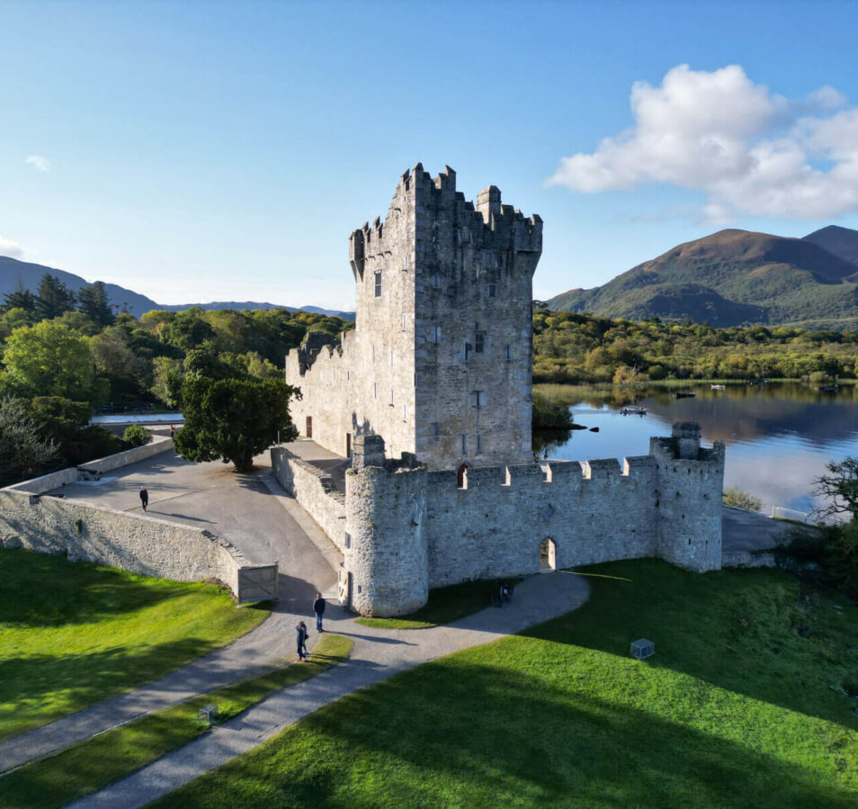 A couple strolls by a historic castle near a serene lake and mountains in Killarney.