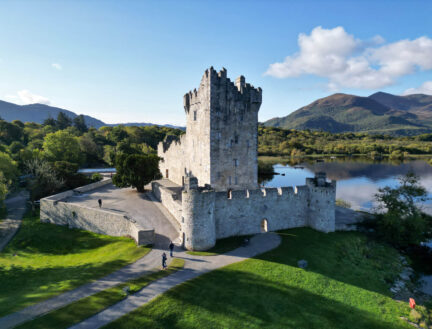 A family walking near a historic stone castle by a calm lake, surrounded by lush greenery and mountains in Killarney.