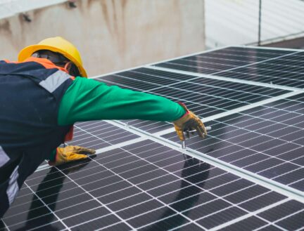 A worker in safety gear installs solar panels on a building roof.