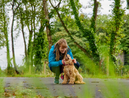 A woman kneels to pet her dog on a path surrounded by lush green trees.