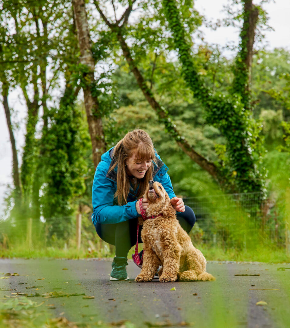 A woman kneels to pet her dog on a path surrounded by lush green trees.