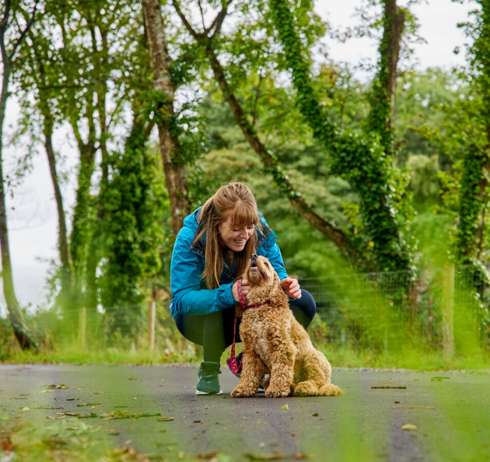 A woman kneels to pet her dog on a path surrounded by lush green trees.